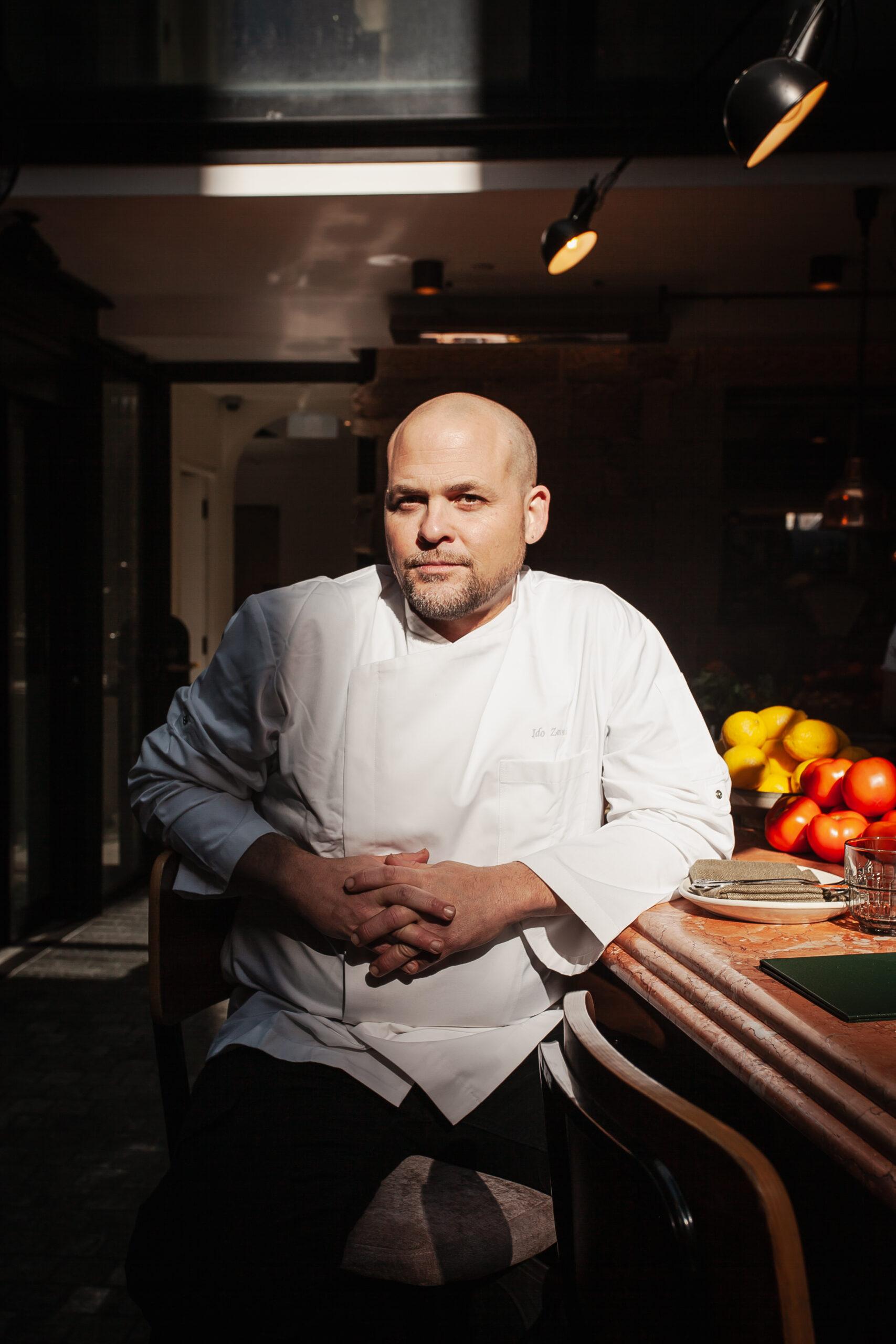 Shaffa Chef Ido Zarmi in white uniform at a marble counter with fresh produce, warm lighting, and elegant restaurant decor.