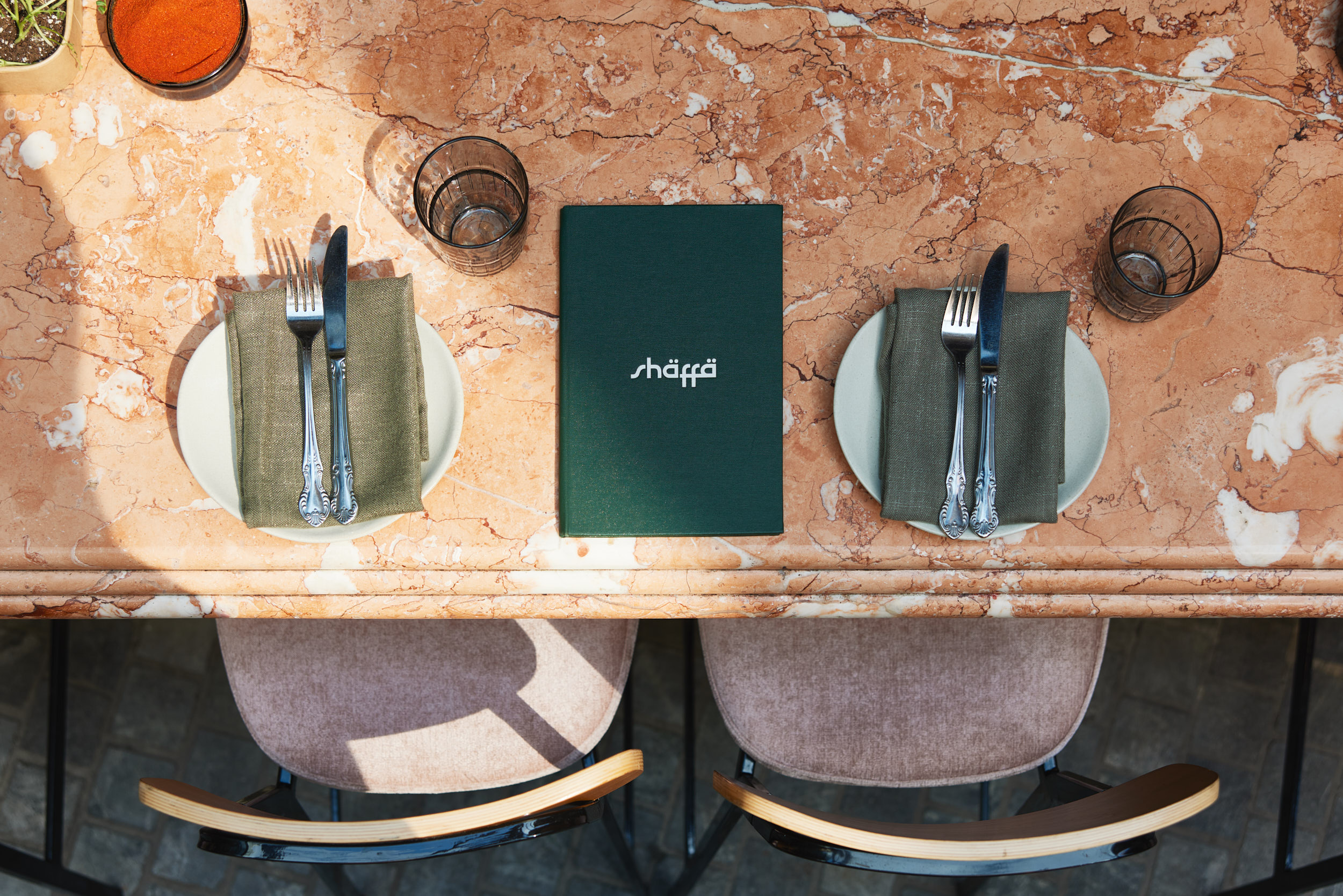 Middle Eastern restaurant chef's table with marble top, 'Shaffa' menu, green napkins, silverware, and velvet chairs set for two.